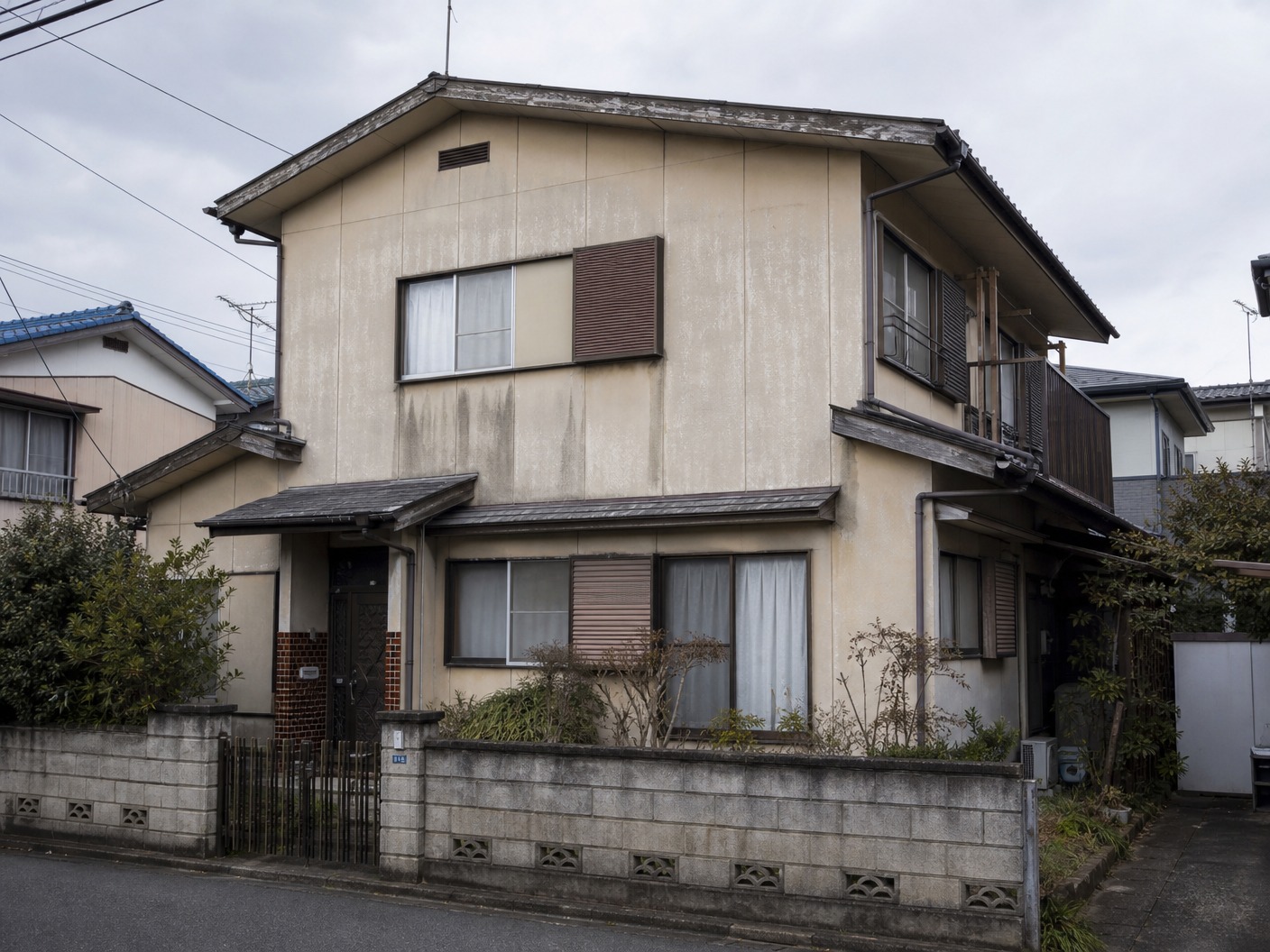 An old Japanese suburban two-story house with fading beige composite siding walls, dirty water stains under the windows, chalking paint surface, worn-out roof appearance.